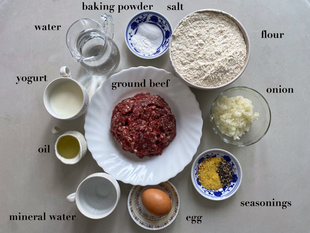 Ten different sized dishes and cups with ingredients  (salt, water, mineral water, oil, yogurt, flour, egg, seasonings, ground beef, onion, salt, et al.), on a gray background.