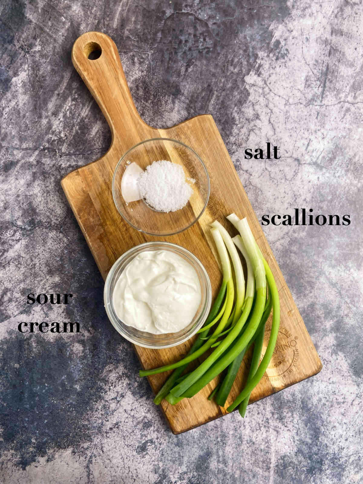 Overhead photo of ingredients (scallions, salt and sour cream) on a cutting board on a marble gray background. 