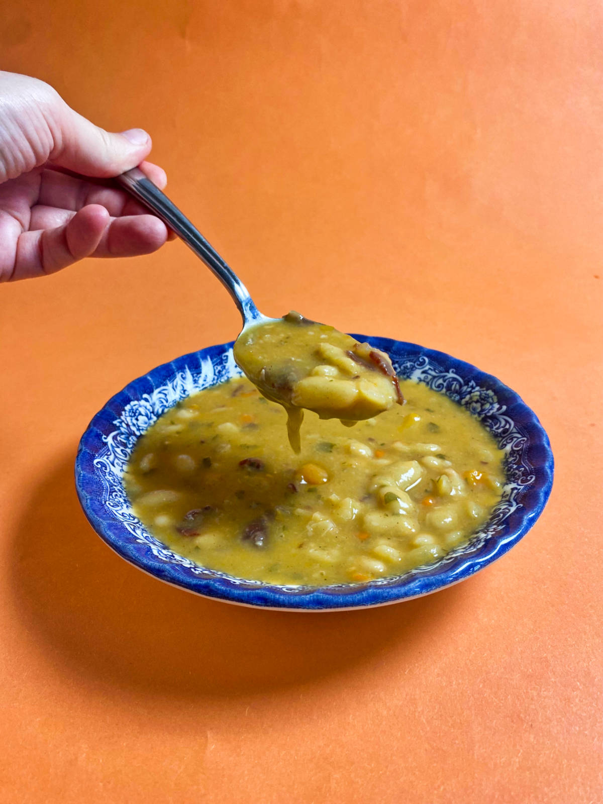 A ladle holding bean soup above the blue bowl, on an orange background.
