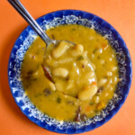 A spoon holding bean soup above the blue bowl, on an orange background.